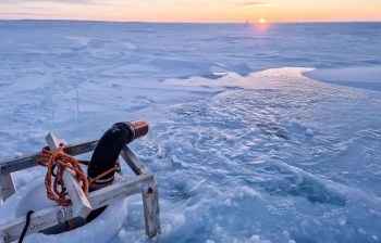 A pipe protrudes from ice, pumping out water under a sunset sky on a snowy landscape.