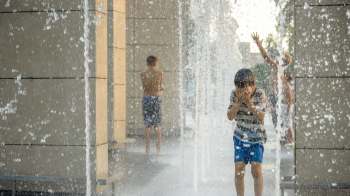 A young boy plays in a fountain, shielding his face from the splashing water while others enjoy the spray nearby.