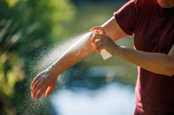 A person sprays insect repellent on their arm in a sunny outdoor setting.