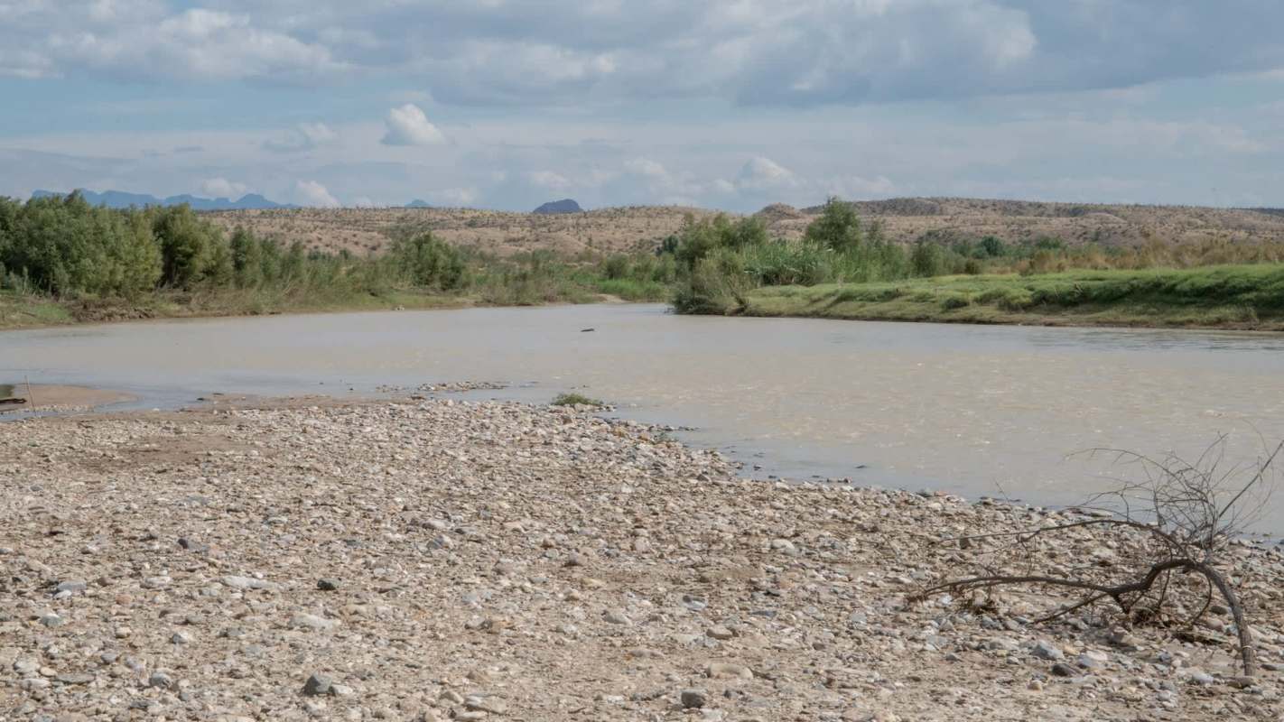 A tranquil riverbank scene with low water levels, pebbles, muddy water, and lush greenery under a partly cloudy sky.