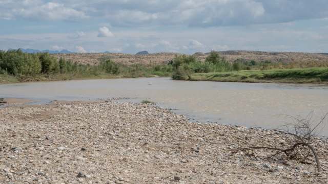 A tranquil riverbank scene with low water levels, pebbles, muddy water, and lush greenery under a partly cloudy sky.