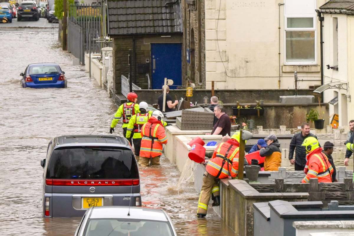 Rescue workers and residents respond to significant flooding, with vehicles submerged and water covering the street.