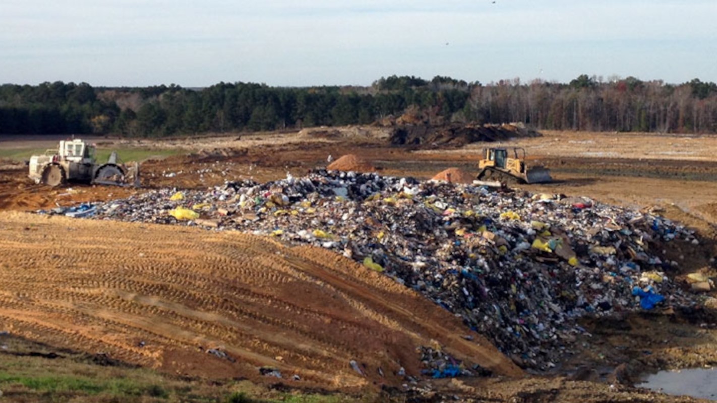 Heavy machinery operates around a large pile of garbage at a landfill under a clear sky.