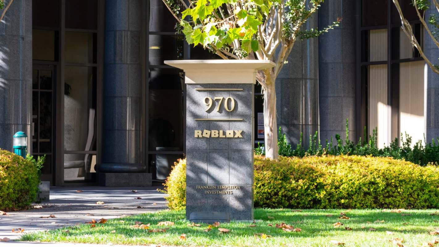 A sign in front of a building displaying the "ROBLOX" logo, surrounded by greenery and a paved walkway.