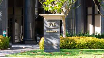A sign in front of a building displaying the "ROBLOX" logo, surrounded by greenery and a paved walkway.