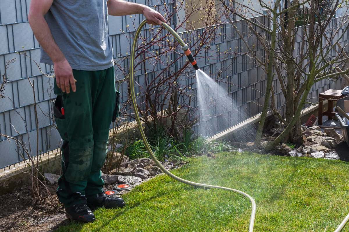A person in a gray shirt waters a lawn with a hose on a sunny day, surrounded by plants and garden furniture.