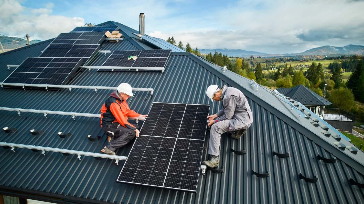 Two technicians install solar panels on a roof, surrounded by scenic mountains and trees under a clear sky.