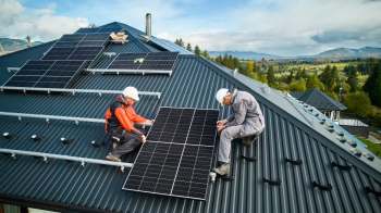 Two technicians install solar panels on a roof, surrounded by scenic mountains and trees under a clear sky.