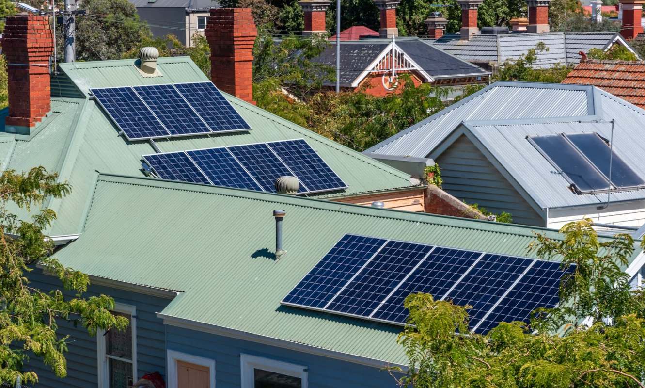 Rooftops with green metal sheeting featuring multiple solar panels under bright sunlight.