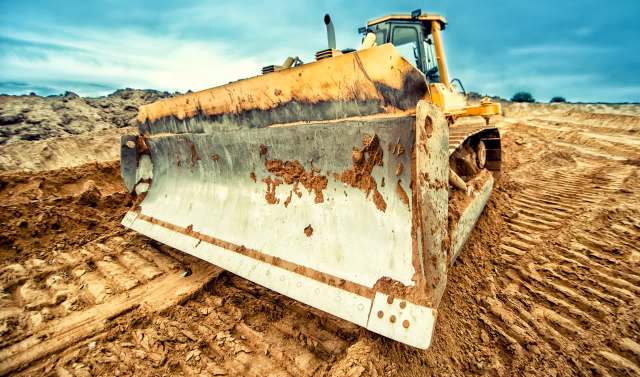 A close-up of a bulldozer's blade on a freshly disturbed dirt surface under a cloudy sky.