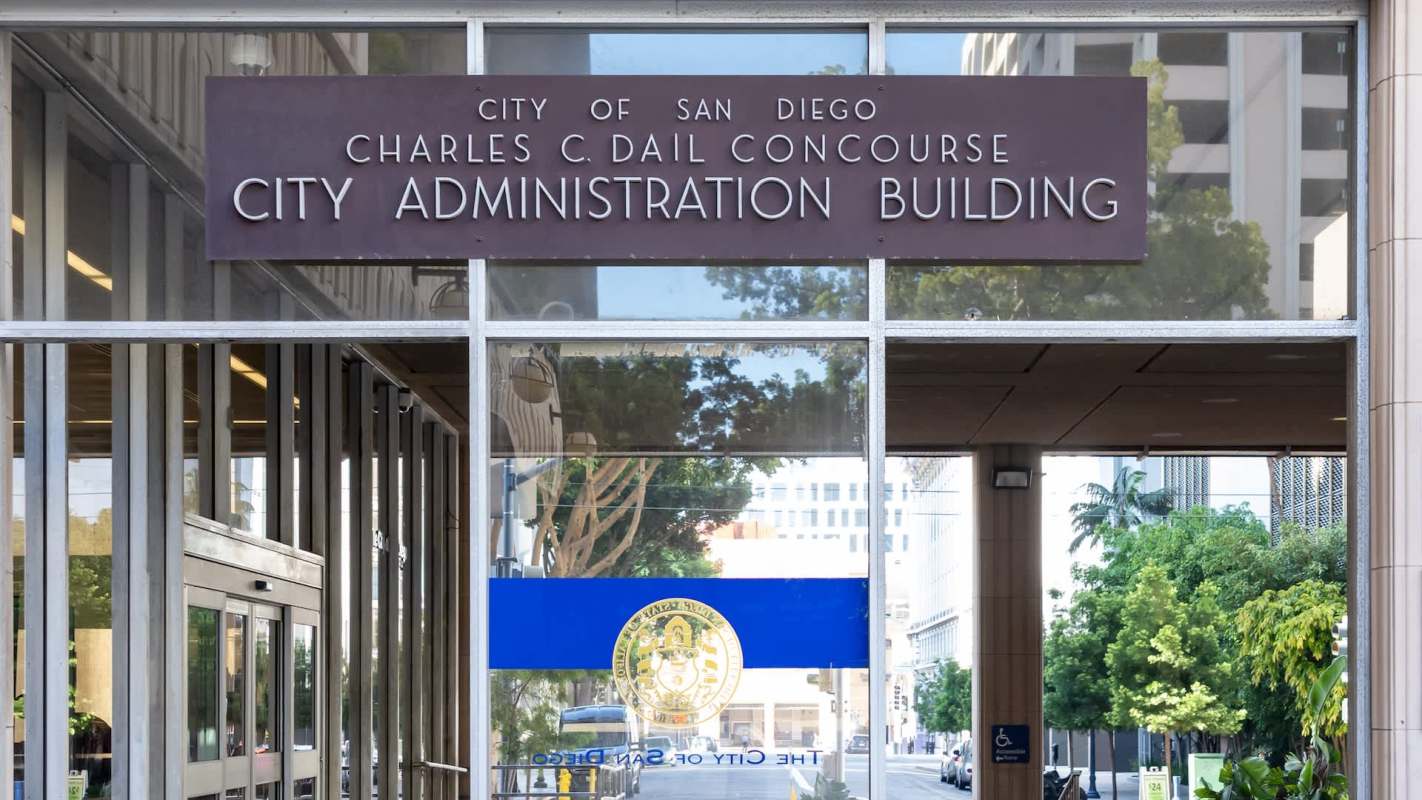 Entrance to the City Administration Building in San Diego, with a sign and glass doors reflecting the surroundings.
