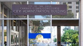 Entrance to the City Administration Building in San Diego, with a sign and glass doors reflecting the surroundings.
