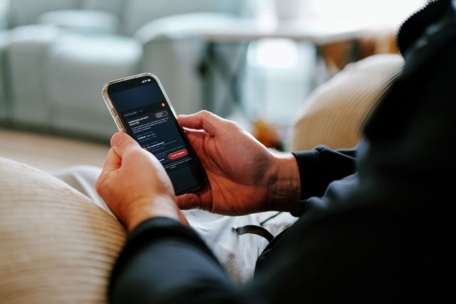 A person sits on a couch, holding a smartphone displaying a screen with a notification about suspicious activity.