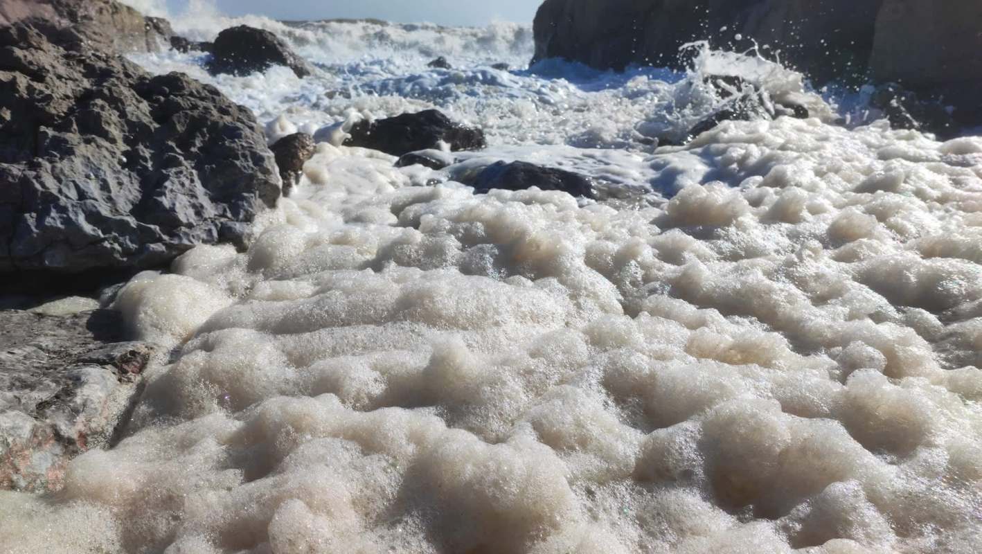 Foamy sea waves crash over rocky shore, creating a bubbly and textured surface against a bright sky.