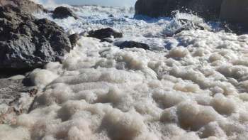 Foamy sea waves crash over rocky shore, creating a bubbly and textured surface against a bright sky.