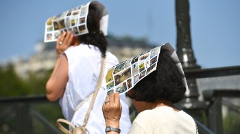 Two people walk outdoors wearing printed paper on their heads for sun protection.