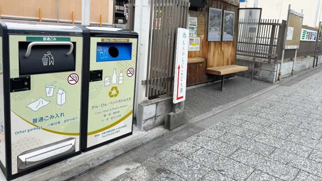 Two bins for waste disposal beside a wooden bench at a bus stop.
