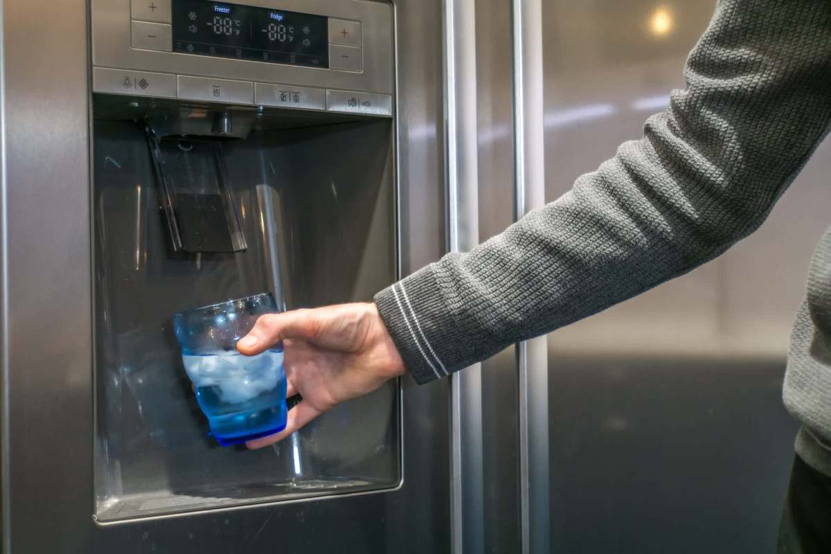 A person fills a blue glass with ice and water from a refrigerator's dispenser.