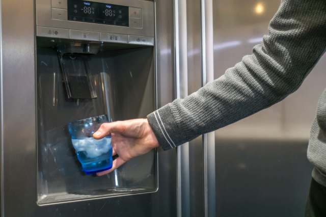 A person fills a blue glass with ice and water from a refrigerator's dispenser.