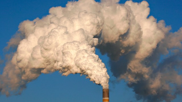 Thick white smoke billows from the top of a tall industrial smokestack against a clear blue sky.