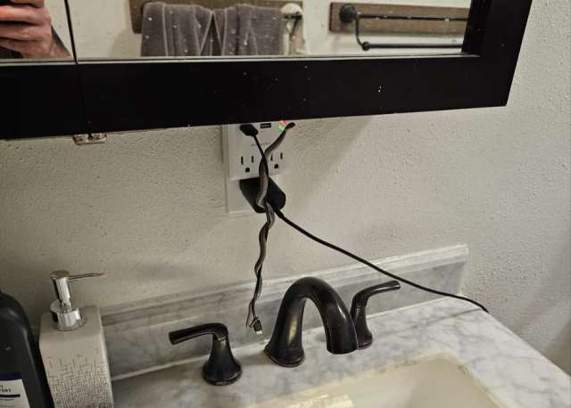 A bathroom countertop featuring a sink, soap dispenser, and a mirror with visible electrical outlet and cords.