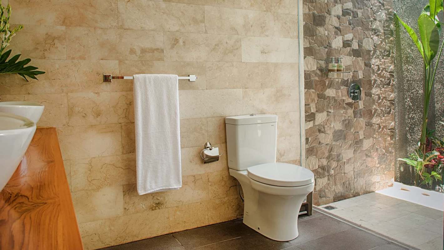 A modern bathroom in a resort featuring a white toilet, towel rack, and stone wall shower with plants visible outside.