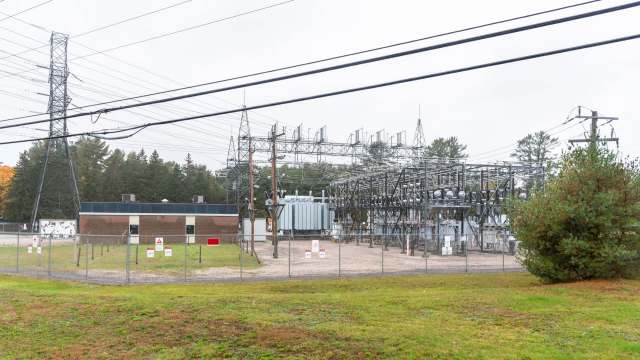 An electrical substation surrounded by a fence, with power lines and a small building in the background.