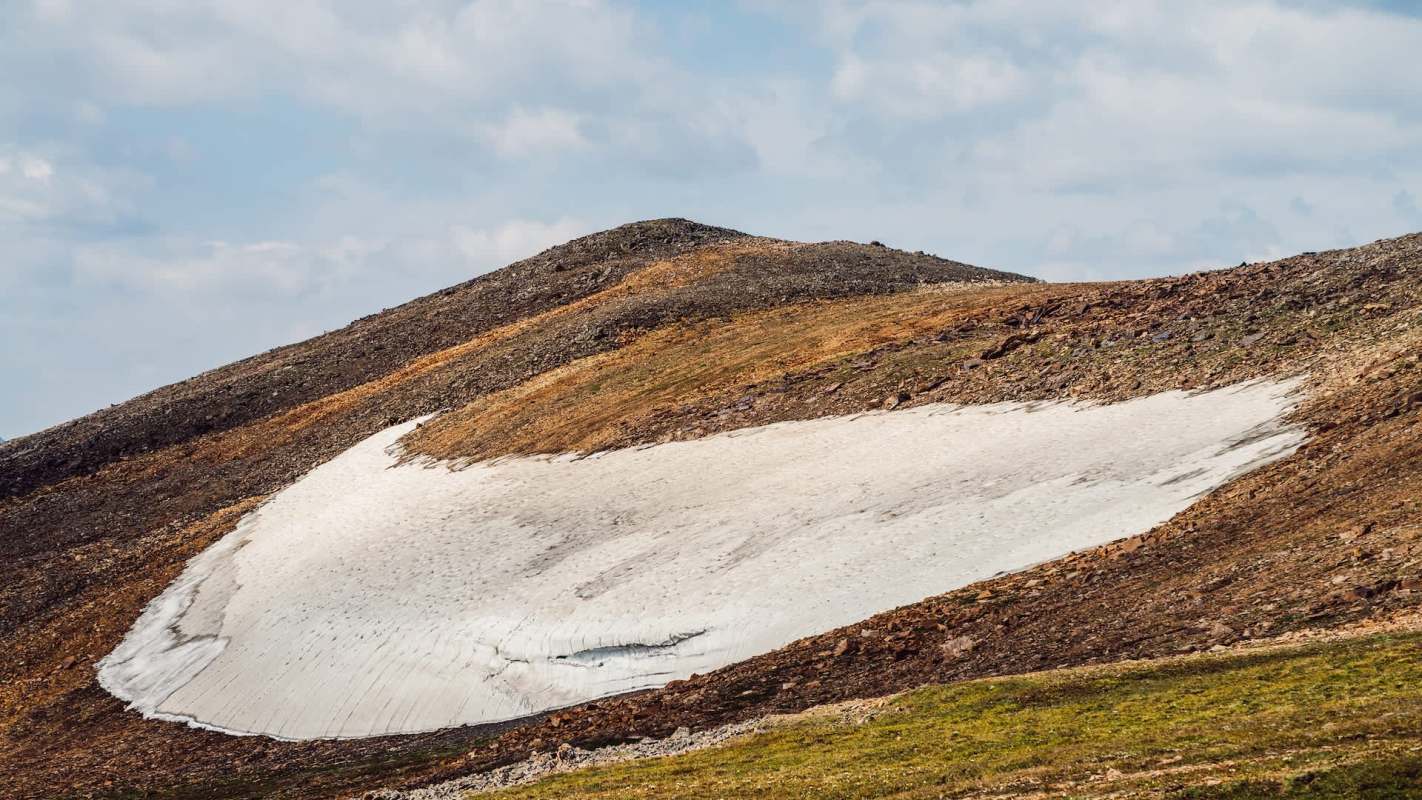 A snow patch rests on a rocky hillside, with scattered green vegetation and a partly cloudy sky in the background.