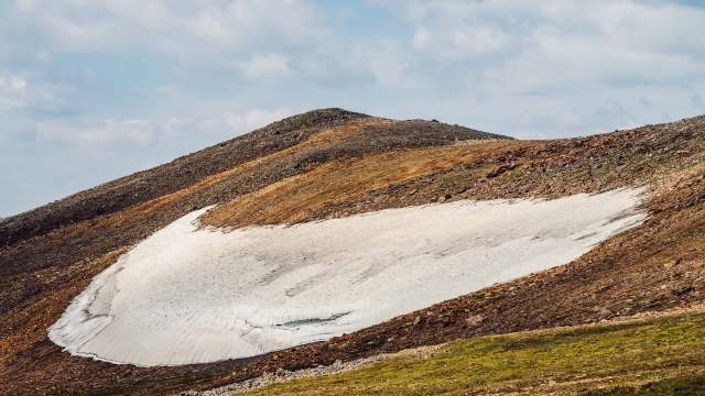 A snow patch rests on a rocky hillside, with scattered green vegetation and a partly cloudy sky in the background.