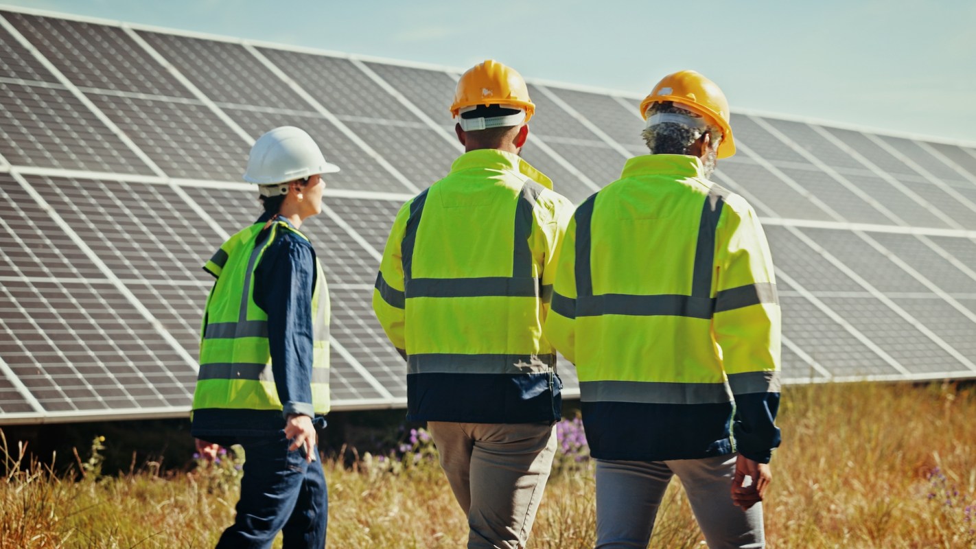 Three workers in safety gear walk past solar panels in a grassy field under a clear sky.