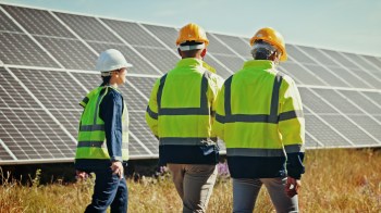 Three workers in safety gear walk past solar panels in a grassy field under a clear sky.