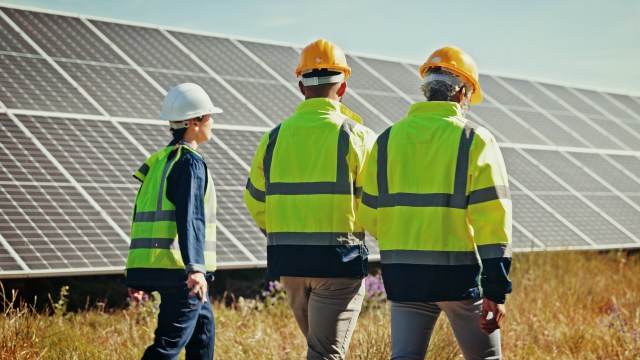 Three workers in safety gear walk past solar panels in a grassy field under a clear sky.