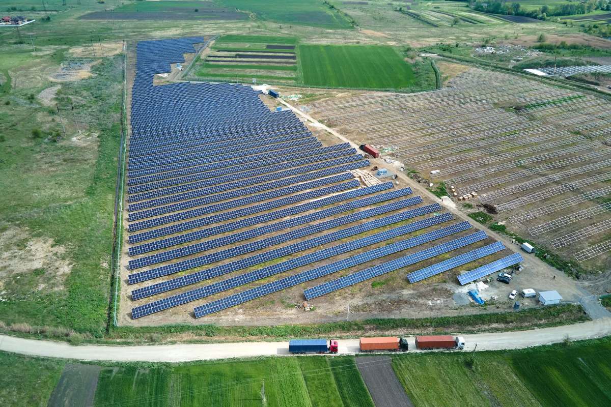 Aerial view of a large solar farm with rows of panels and trucks on a dirt road nearby.