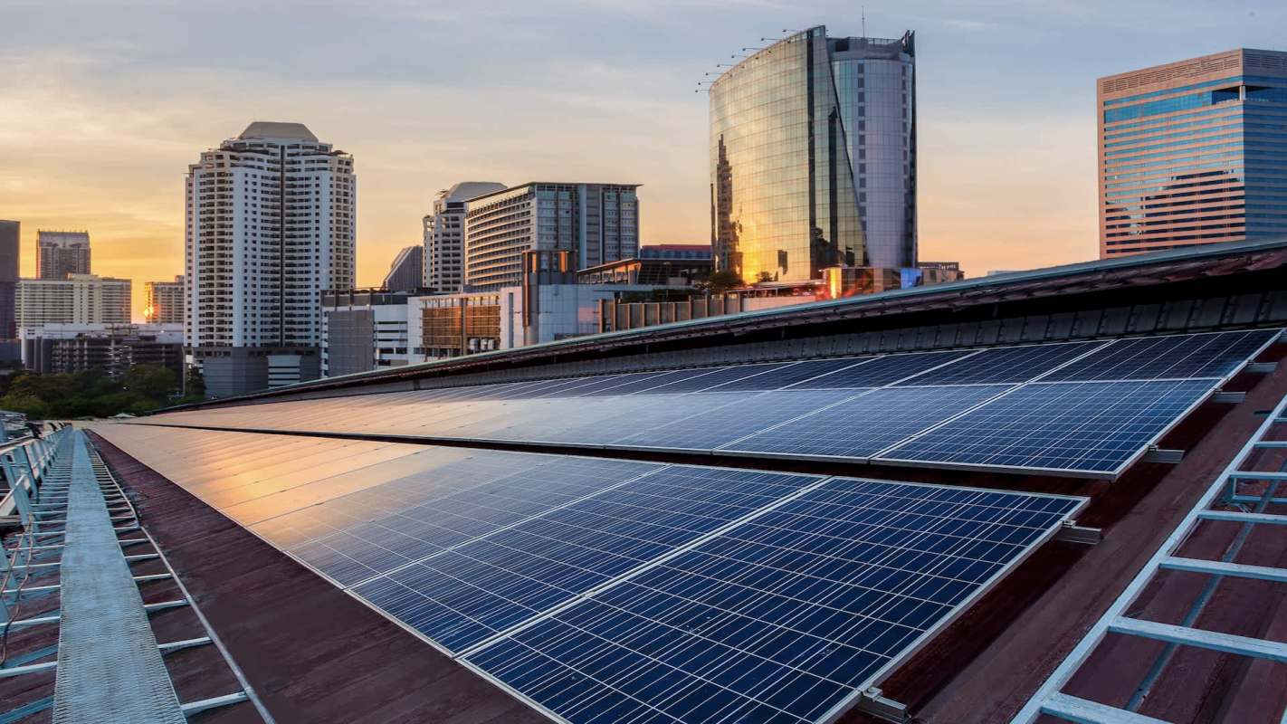A rooftop covered with solar panels with a city skyline and sunset in the background.