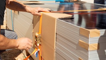 A person tightening a strap around a stack of solar panel boxes on a construction site.