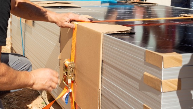 A person tightening a strap around a stack of solar panel boxes on a construction site.