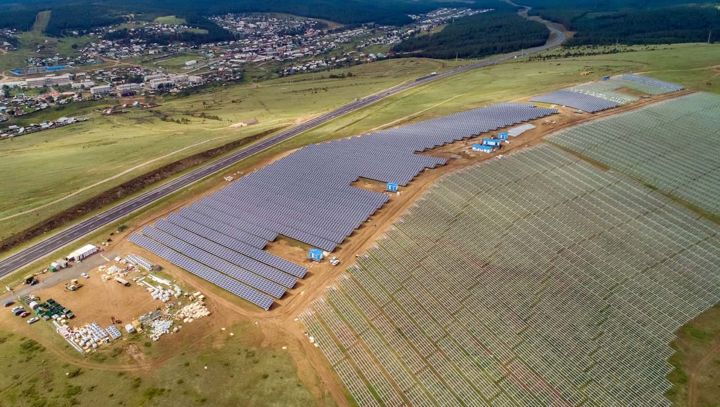 An aerial view of a large solar farm with rows of solar panels and a nearby town in the background.