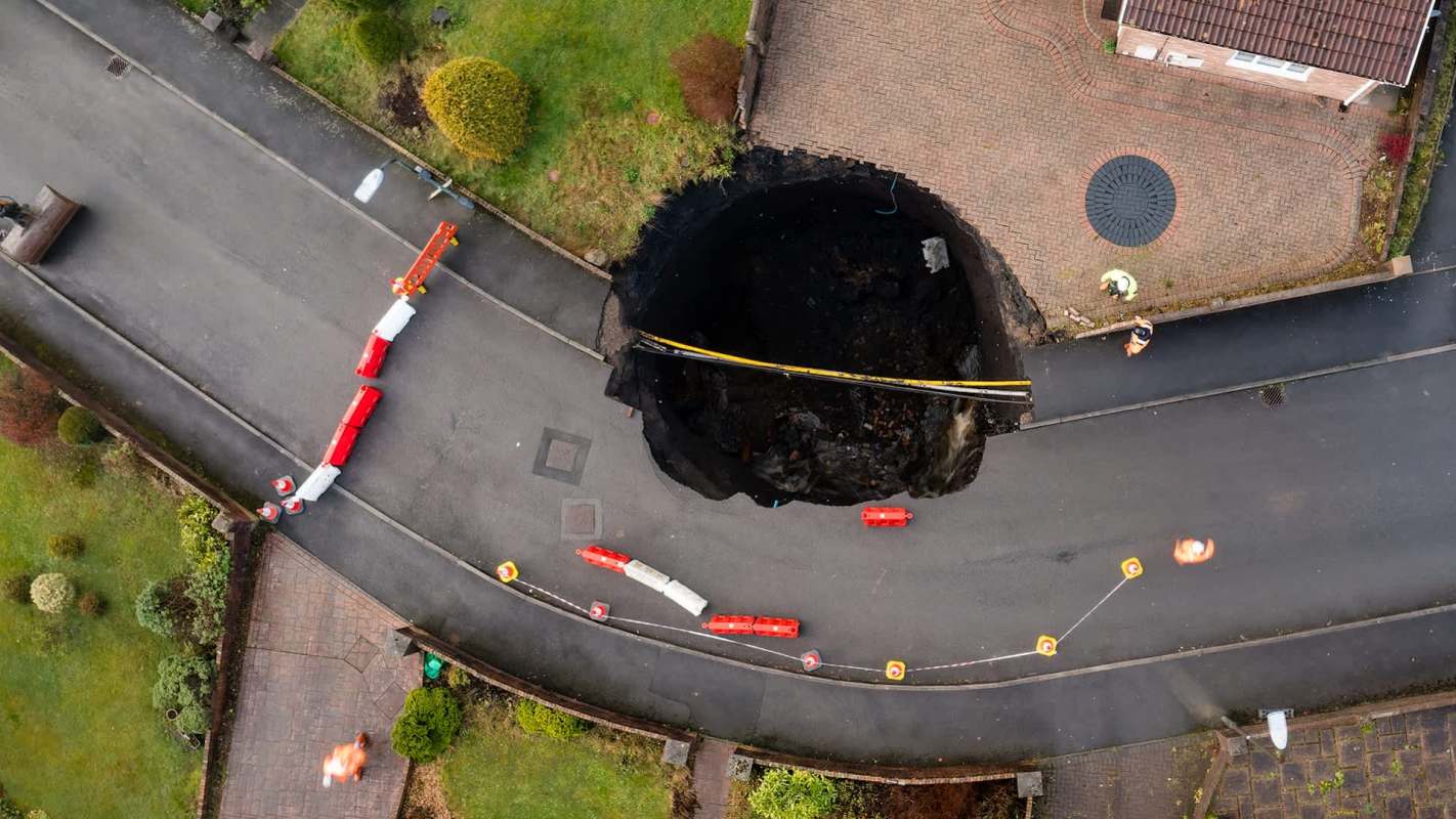 A large sinkhole on a residential road, with traffic barriers in place and construction workers surveying the damage.
