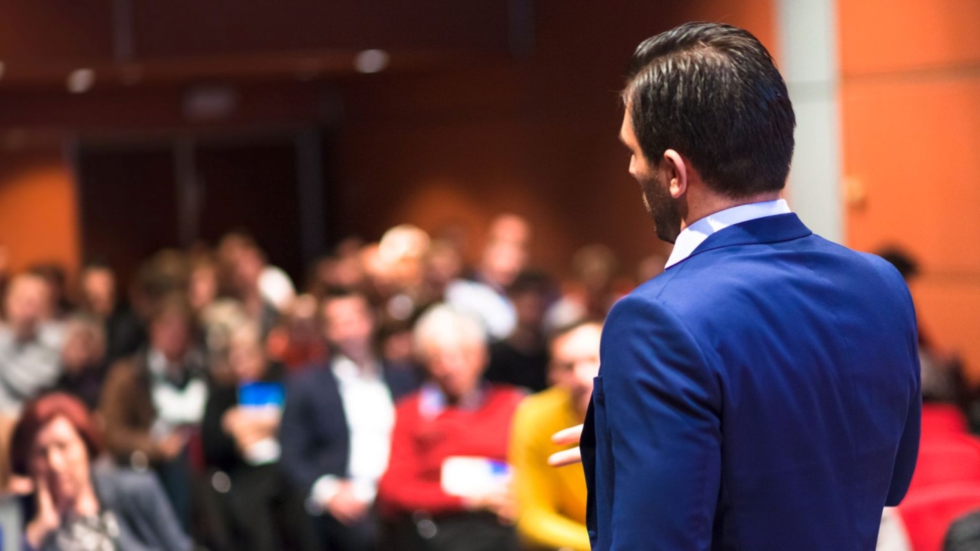 A man speaks to a crowd of attentive listeners in a meeting setting.