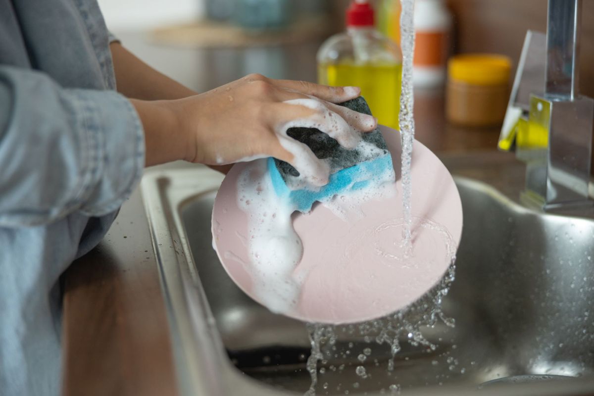 A person washes a pink plate with a soapy sponge under running water at a kitchen sink.