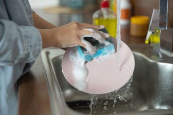 A person washes a pink plate with a soapy sponge under running water at a kitchen sink.