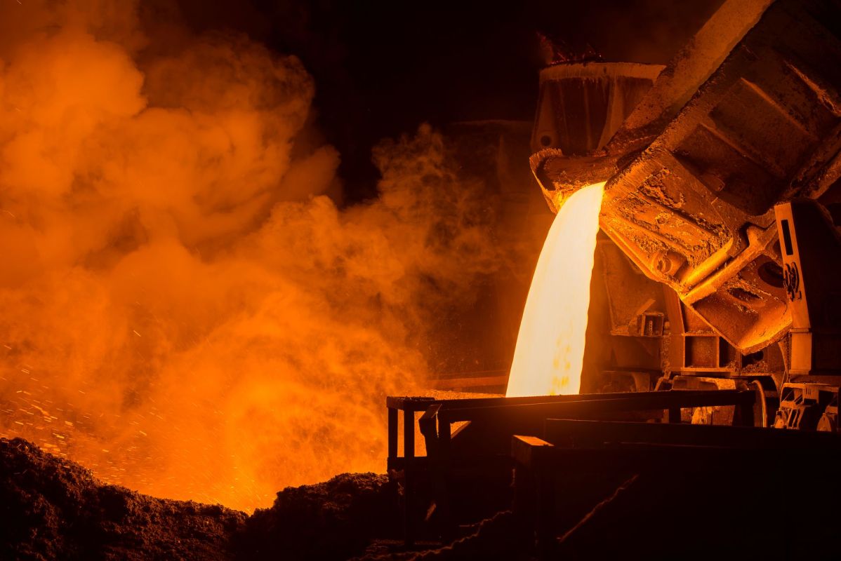 Molten metal pours from a furnace, surrounded by glowing orange smoke and sparks in a steel factory.