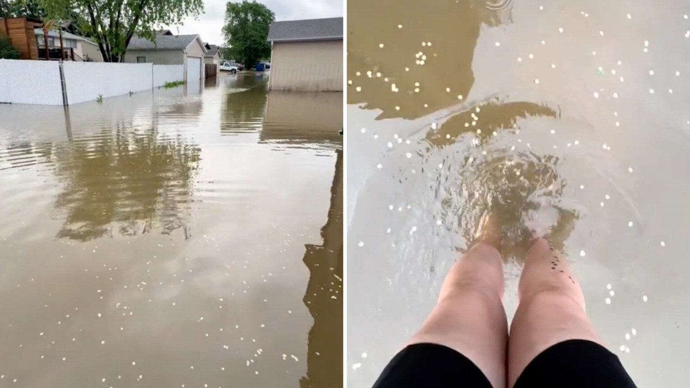 A flooded street with water reflecting nearby trees and a person wading through the shallow water with bare legs.