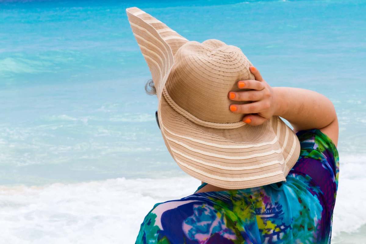 A woman in a colorful, patterned cover-up and a wide-brimmed hat gazes at the ocean.