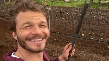 A smiling man holding a gardening tool stands in a garden with soil and plants in the background.