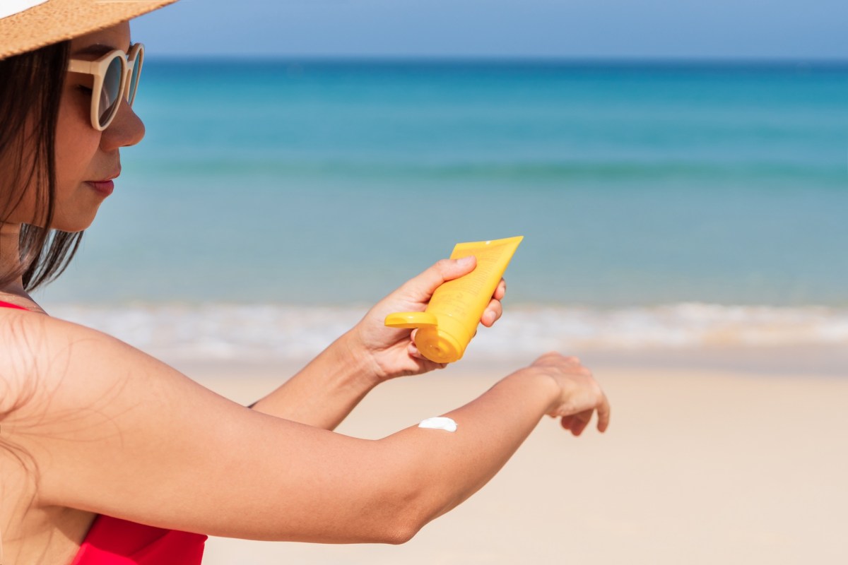 A woman in sunglasses applies sunscreen to her arm at the beach with the ocean in the background.