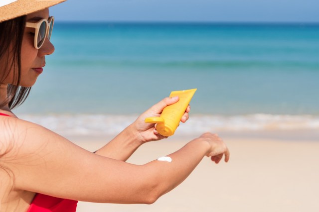 A woman in sunglasses applies sunscreen to her arm at the beach with the ocean in the background.