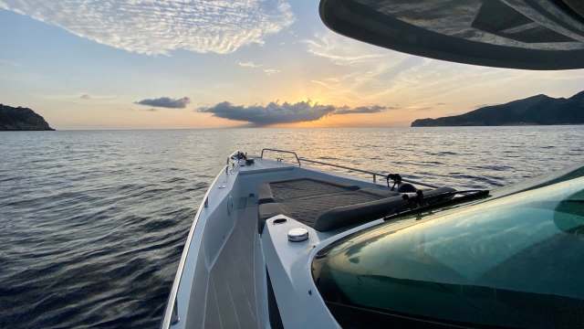 A view from the bow of a boat overlooking calm waters at sunset, with distant hills and clouds on the horizon.