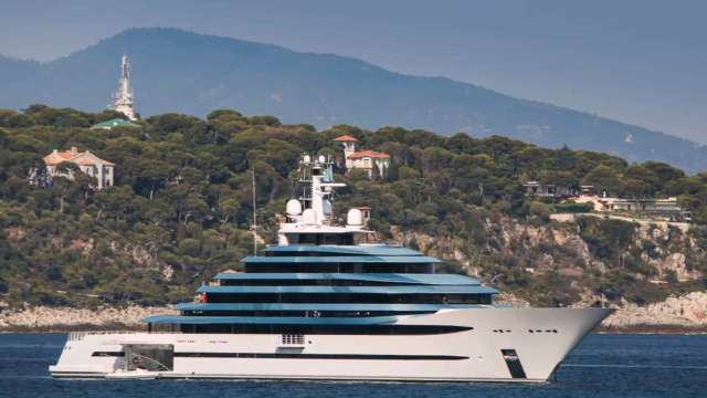 A luxurious yacht sails past a lush green hillside with elegant homes and towering mountains in the background.