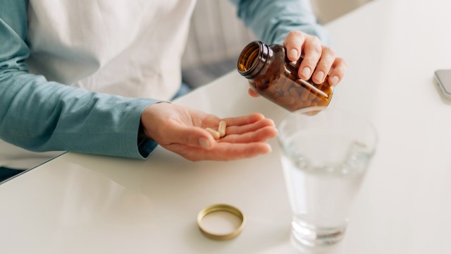 A person pours capsules from a brown bottle into their palm, with a glass of water nearby.